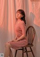 A woman sitting on a wooden chair in a pink dress.