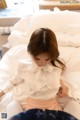 A woman sitting on a bed wearing a white blouse and pink skirt.