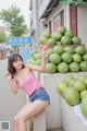 A woman in a pink tank top and denim shorts posing in front of a pile of watermelons.